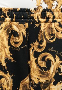 Κοντινό πλάνο μαύρου υφάσματος με χρυσά μπαρόκ floral σχέδια και την επιγραφή "Versace Jeans", που παρουσιάζει μια ελαστική ζώνη μέσης.