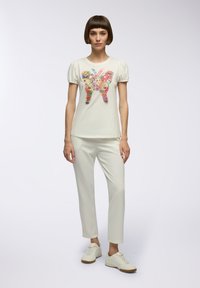 T-shirt crema con grafica di una farfalla colorata, maniche a palloncino corte, abbinata a pantaloni crema e sneakers chiare. Vestibilità semplice e moderna.