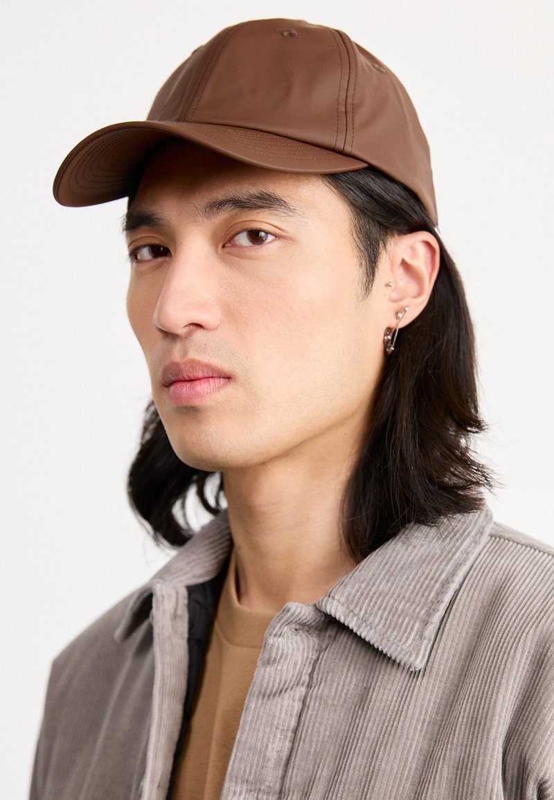 Casquette de baseball en cuir brun avec une visière cousue, portée par une personne dans une veste en velours côtelé grise texturée, avec une chemise beige en dessous.