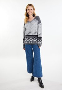 Grau und schwarz gemusterter Strickpullover mit V-Ausschnitt, der ein Zickzackdesign aufweist, getragen mit weit geschnittenen blauen Jeans und schwarzen Stiefeletten.
