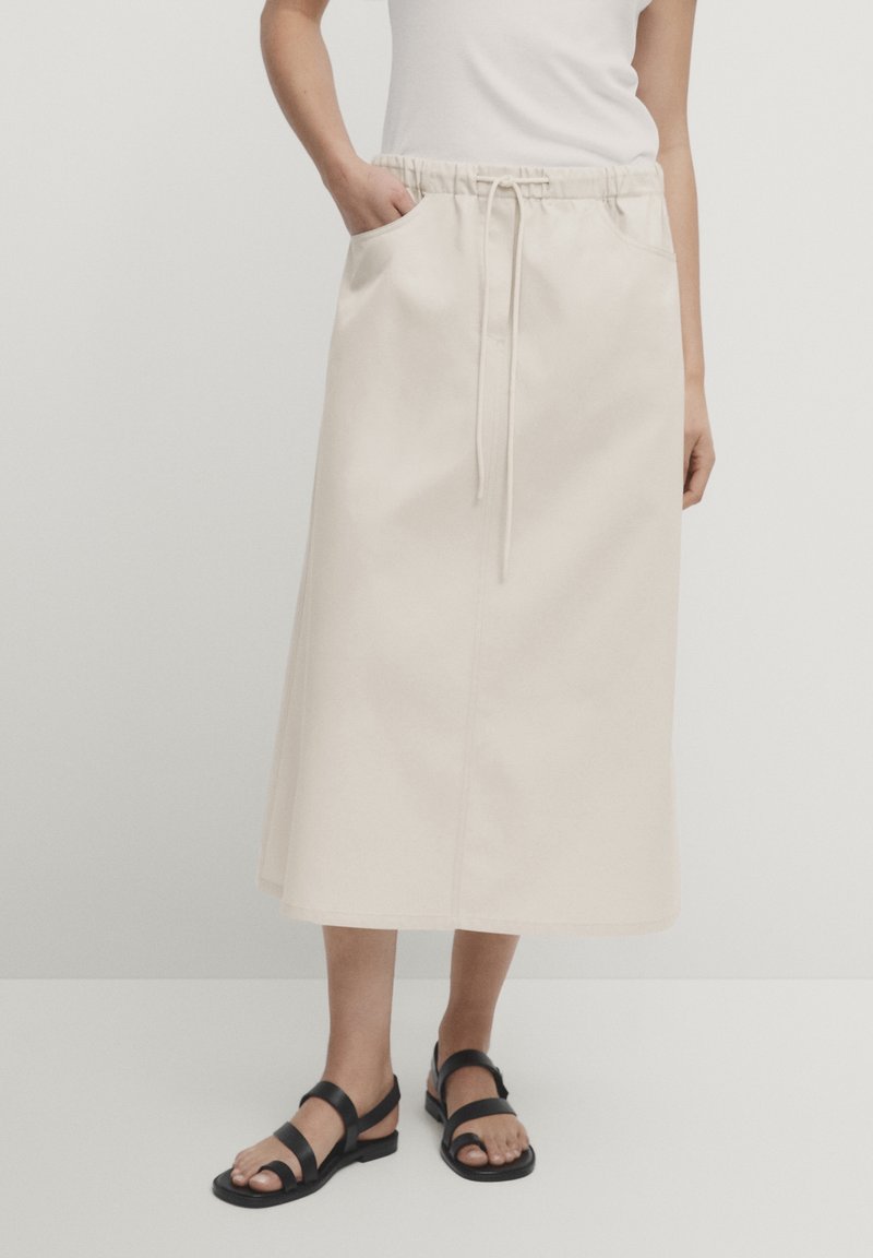 Femme portant une jupe midi crème avec taille à cordon, haut blanc et sandales plates noires, debout avec une main dans la poche.