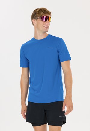 Junger Mann trägt blaues Sport-T-Shirt und schwarze Shorts mit verspiegelten Sportsonnenbrillen, steht und schaut zur Seite, lächelt.