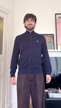 Muž s tmavými vlasmi a bradou stojí v interiéri, oblečený v tmavomodrom svetri na zips a tmavých nohaviciach, na pozadí sú farebné obrazy v rámikoch.