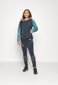 Σετ επαλειμμένων ρούχων εξωτερικού χώρου που περιλαμβάνει μια μαύρη κεντητής ζακέτα με τιρκουάζ μανίκια και κοραλί κουκούλα, σε συνδυασμό με ναυτικό κολάν και μαύρα παπούτσια.