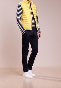 Gilet rembourré jaune sur une chemise à rayures bleu marine à manches longues, associé à un pantalon foncé et des baskets blanches. Tissu lisse, design ajusté.