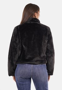 Giacca nera in pelliccia sintetica con colletto alto, maniche lunghe e vestibilità ampia, abbinata a jeans in denim blu. Tessuto morbido e senza chiusure visibili.