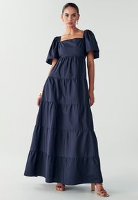 Navy blue maxi mekko kevyestä kankaasta, jossa on neliönmuotoinen kaula-aukko, puffihihat, kerroksellinen muotoilu ja sileä rakenne.