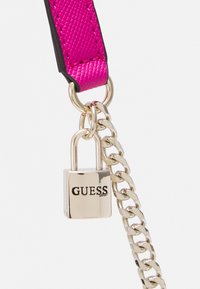 Un lucchetto color oro con la scritta "GUESS" incisa, attaccato a una catena dorata e a una striscia testurizzata rosa con bordi neri.