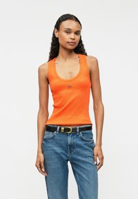 Narancssárga bordázott ujjatlan fels�ő, kerek nyakkivágással, kis kék logóval. Kék farmernadrággal és fekete övvel, amelynek arany díszítése van párosítva.