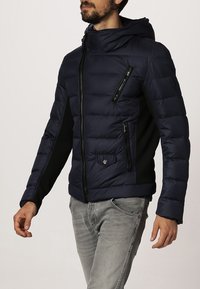 Marinblå puffjacka med huva, svart accent, kviltad struktur, dragkedjefickor och en figurnära design.