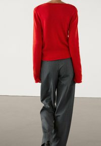 Personne portant un pull rouge à manches longues et un pantalon gris lâche, se tenant contre un mur blanc uni et un sol gris.