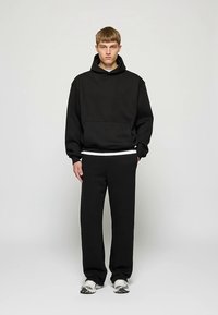 Musta puuvillainen huppari, jossa on suuri etutasku, yhdistettynä vastaaviin mustiin collegehousuihin. Molemmissa on rento istuvuus ja pehmeä tunnelma.