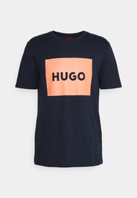 Námorně modré tričko s krátkým rukávem vyrobené z bavlny s velkým broskvovým čtvercovým logem se slovem "HUGO" napsaným výraznými černými písmeny. Hladká textura, kulatý výstřih.