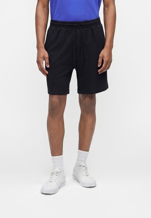 Άνδρας που φορεί μαύρα υφασμάτινα σορτς με κορδόνι, λευκές κάλτσες και λευκά αθλητικά παπούτσια, στέκεται μπροστά σε απλό ανοιχτογκρι φόντο.