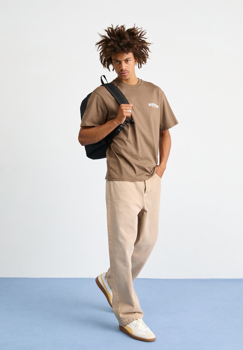 Καφέ κοντομάνικο μπλουζάκι με λευκό λογότυπο, μπεζ παντελόνι και λευκά αθλητικά παπούτσια με κίτρινες λεπτομέρειες. Μαύρος σακίδιο προσθέτει λεπτομέρεια.