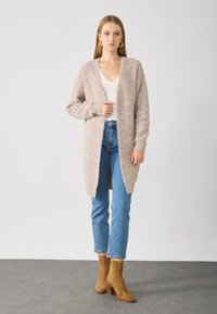 Cardigan rose pâle, fluffy, à ouverture frontale, manches longues et poignets côtelés. Associé à un jean droit bleu et des bottes à talons marron.