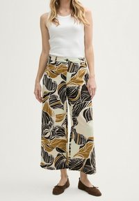 Kobieta w szerokich spodniach w odważne brązowe i czarne wzory liści, w białej bezrękawnikowej bluzce i brązowych płaskich butach, stojąca na tle jednolitego tła.