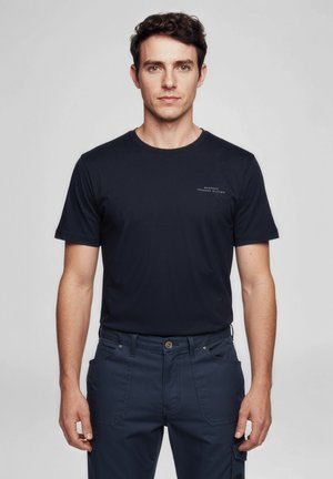 Junger Mann mit kurzen lockigen Haaren, der ein marineblaues kurzärmliges Hemd und marineblaue Hosen trägt, vor einem einfarbigen, hellen Hintergrund stehend.