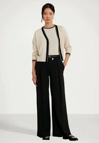 Sieviete, kas ģērbusies krēmkrāsas kardiganā ar melniem akcentiem, krēmkrāsas blūzē, melnās plašās biksēs un melnos loafers, stāvot uz fona ar vienkāršu baltu krāsu.