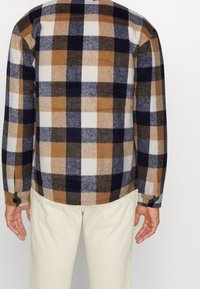 Brun, marinblå och vit rutigt skjorta med knappar och långa ärmar samt sidofickor. Tillverkad av ett mjukt, strukturerat tyg. Detalj med två knappar vid manschetterna.