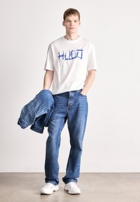 Tricou alb cu text grafic albastru, asociat cu blugi largi din denim albastru și o geacă din denim rayée albastră, completat cu adidași albi.