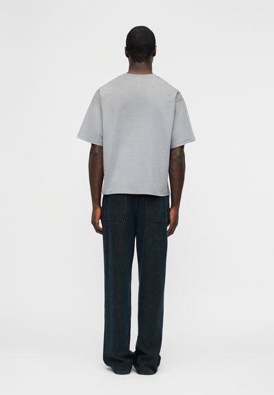 Άνδρας με την πλάτη, φοράει ένα άνετο ανοιχτό γκρι κοντομάνικο μπλουζάκι και σκούρα τζιν φαρδιά στο κάτω μέρος, μπροστά σε απλό φόντο.