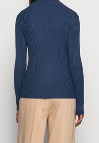 Námořnický žebrovaný pletený svetr s dlouhými rukávy a vysokým výstřihem, s texturovaným povrchem a přizpůsobeným střihem. Nosený s světle barevnými kalhotami.