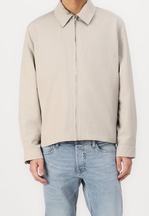 Jasno beżowa kurtka z kołnierzykiem, zamkiem z przodu i bocznymi kieszeniami. Tkanina wydaje się gładka, z nieco dopasowanym krojem.