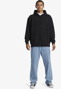 Zwarte hoodie met een kangoeroezak, capuchon met trekkoord, gedragen over lichtblauwe jeans. Zwart-witte sneakers maken de outfit compleet.