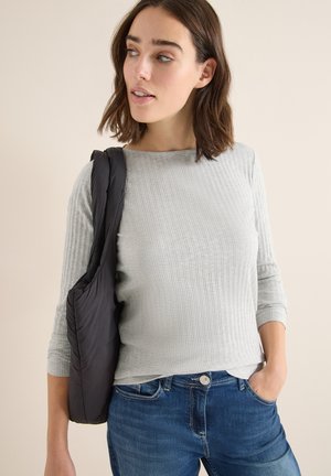 Grijze geribbelde langemouwtop gecombineerd met blauwe jeans; model draagt een zwarte gewatteerde tas over één schouder. Eenvoudig, gestructureerd design.