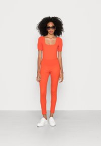 Macacão laranja com decote quadrado e mangas bufantes. Leggings texturizadas, combinadas com ténis brancos. Óculos de sol completam o visual.