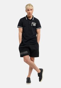 Sort polo med hvide detaljer, logo på brystet; parret med sorte shorts med grafisk tekst. Footwear er sorte med en hvid sål.