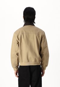 Chaqueta bomber de algodón color caqui con cuello marrón, puños y dobladillo elásticos; presenta una textura suave y costuras traseras. Se llevan pantalones negros debajo.