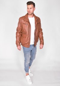 Brun läderjacka med dragkedjefickor, ståkrage och mjuk textur, bärs över en vit t-shirt och kombineras med slitna blå jeans.