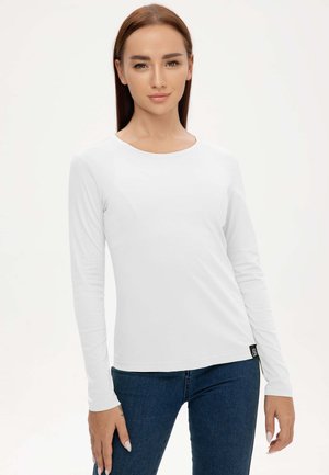 Frau mit glatten braunen Haaren trägt ein schlichtes weißes Langarmshirt und blaue Jeans, steht vor einem hellen Hintergrund.