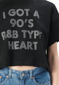 Juodas trumpas medvilninis marškinėliai, puošti sidabriniais rhinestone užrašais: "AŠ TURIU 90-ųjų R&B ŠIRDĮ." Dėvimi su šviesiai mėlynais aukštu liemeniu džinsais.