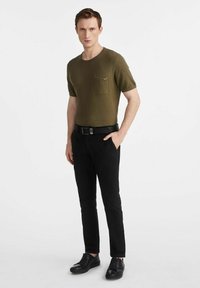 Mężczyzna stoi pewnie, ubrany w oliwkową koszulę z krótkim rękawem, czarne spodnie i czarne buty, na minimalistycznym tle.