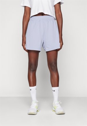 Personne portant un short de sport violet clair, un t-shirt court blanc, des chaussettes Nike blanches et des chaussures de course blanches et jaunes, debout sur un fond uni.