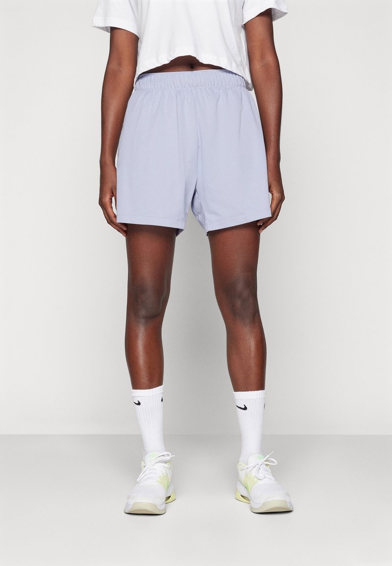 Personne portant un short de sport violet clair, un t-shirt court blanc, des chaussettes Nike blanches et des chaussures de course blanches et jaunes, debout sur un fond uni.
