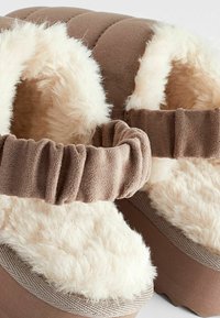 Chaussons marron en tissu doux, avec une doublure crème duveteuse, une bride élastique et des semelles texturées pour une meilleure adhérence.