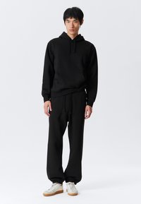 Haut à capuche noir avec une poche frontale et un cordon de serrage, associé à un pantalon de survêtement noir avec une taille élastique et des poignets, porté avec des baskets blanches.