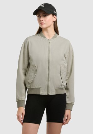 Femeie purtând o jachetă bomber gri deschis cu fermoar, pantaloni scurți negri de ciclism și o șapcă neagră de baseball cu un logo alb, stând în picioare pe un fundal simplu.