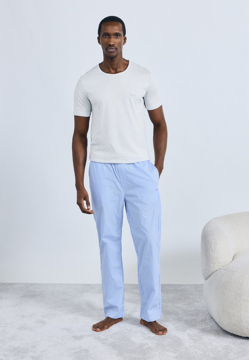 Svetlo siva prilegajoča majica s kratkimi rokavi, kombinirana z modrimi in belimi karo pižamastimi hlačami. Model stoji boso na mehkem preprogi.