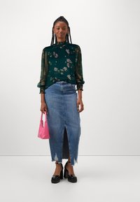 Blusa floreale verde con maniche trasparenti, gonna di jeans con spacco frontale e orlo sfilacciato, scarpe nere con plateau, mentre tiene in mano una piccola borsa rosa.