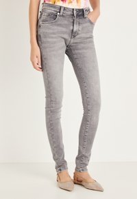 Szare obcisłe dżinsy z wysokim stanem o miękkiej fakturze, z lekkim blaknięciem i pięcioma kieszeniami; noszone z beżowymi wsuwanymi butami z plecionym wzorem.