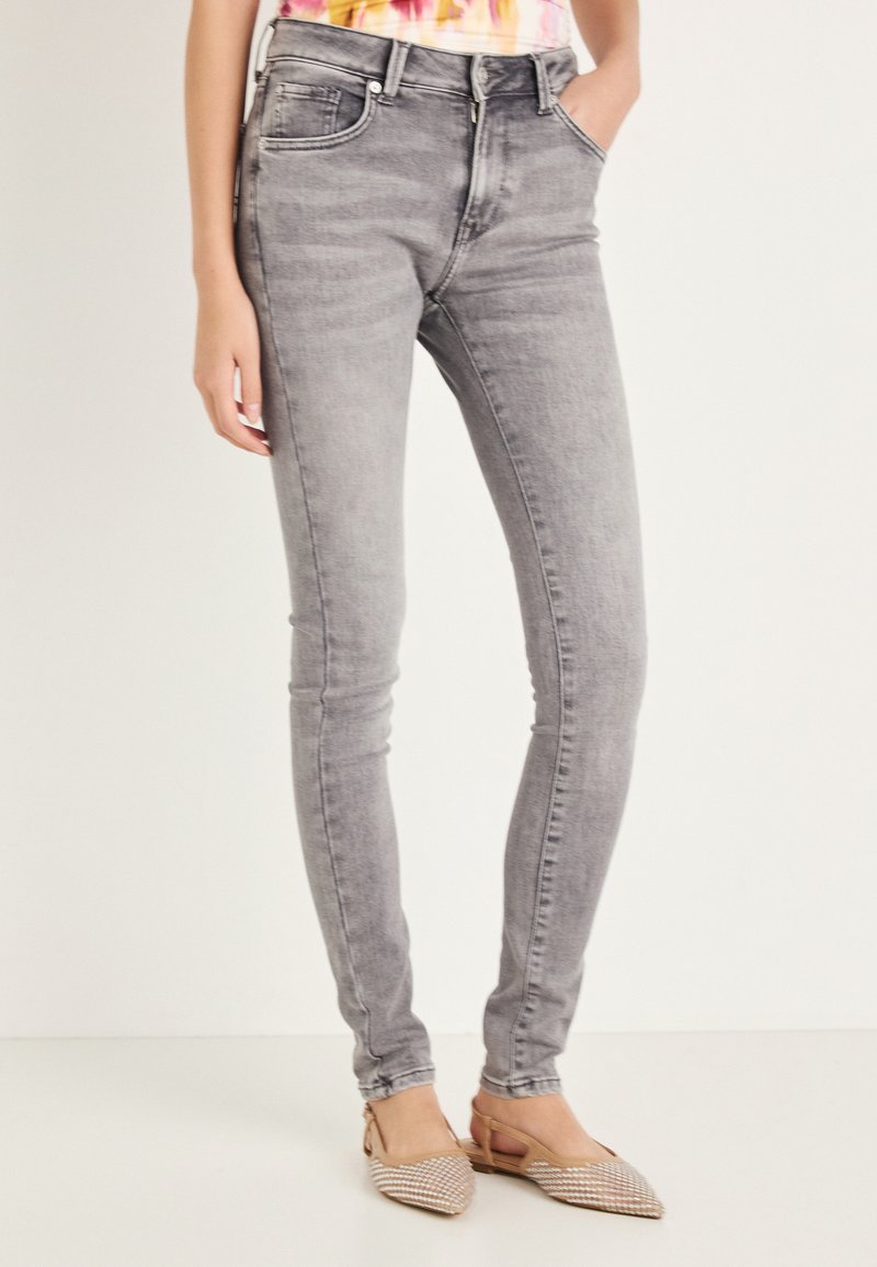 Szare obcisłe dżinsy z wysokim stanem o miękkiej fakturze, z lekkim blaknięciem i pięcioma kieszeniami; noszone z beżowymi wsuwanymi butami z plecionym wzorem.