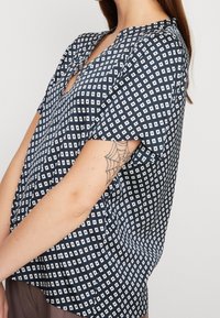 Top blu navy con maniche corte, caratterizzato da un motivo geometrico a quadrati bianchi. Tessuto leggero, vestibilità comoda e scollatura rotonda.