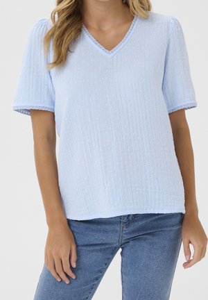 Mujer que lleva una blusa de manga corta texturizada azul claro con ribete de encaje y vaqueros azules, de pie con los brazos relajados a los lados.