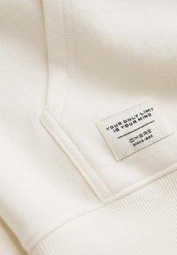 Witte sweatshirtstof met een gestructureerd oppervlak. Bevat een label met zwarte tekst waarop staat "JE ENIGE BEPERKING IS JE GEEST" en de merknaam "OMBRA."