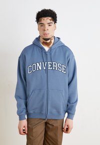 Felpa con zip di colore blu con il testo "CONVERSE", dotata di cappuccio, tasche frontali e polsini a coste. Il materiale sembra morbido e testurizzato.